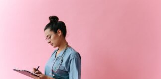 Female nurse in scrubs writing on a clipboard with a stethoscope on a pink background.