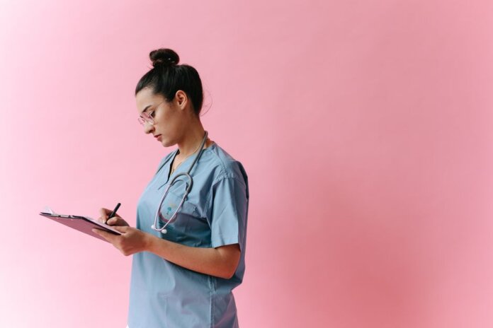 Female nurse in scrubs writing on a clipboard with a stethoscope on a pink background. Female nurse in scrubs writing on a clipboard with a stethoscope on a pink background.