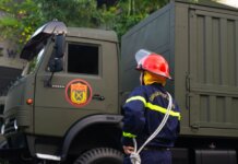 Firefighter in uniform with protective gear stands by an army truck outdoors.