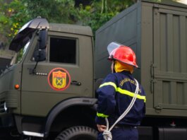 Firefighter in uniform with protective gear stands by an army truck outdoors.