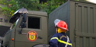 Firefighter in uniform with protective gear stands by an army truck outdoors.