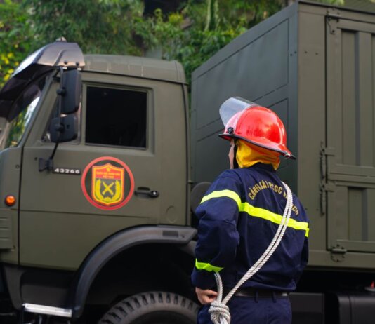 Firefighter in uniform with protective gear stands by an army truck outdoors.