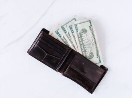 From above of black leather wallet with pockets filled with heap of dollar banknotes on white table