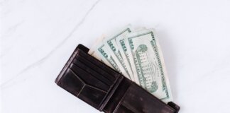 From above of black leather wallet with pockets filled with heap of dollar banknotes on white table