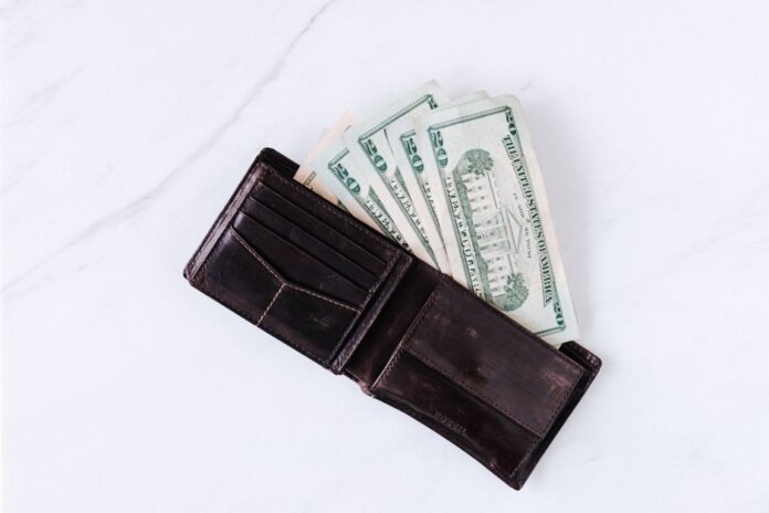 From above of black leather wallet with pockets filled with heap of dollar banknotes on white table From above of black leather wallet with pockets filled with heap of dollar banknotes on white table