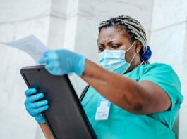 Low angle of focused black female nurse in mask and gloves standing in hospital and looking through papers