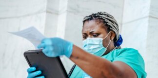 Low angle of focused black female nurse in mask and gloves standing in hospital and looking through papers