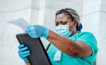 Low angle of focused black female nurse in mask and gloves standing in hospital and looking through papers