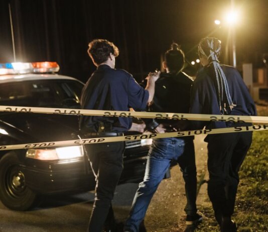 Nighttime arrest scene with police officers and patrol car behind caution tape.