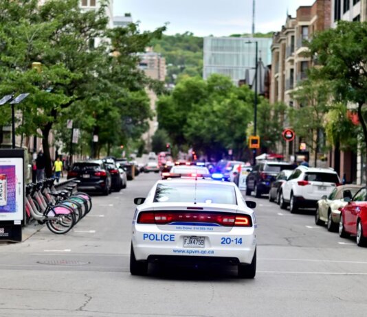 Police car driving through urban street lined with trees and parked bicycles.