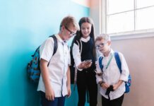 Three children in school uniforms using a smartphone, standing near a blue wall indoors.