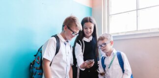 Three children in school uniforms using a smartphone, standing near a blue wall indoors.