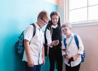 Instagram расширяет программу для школ: новые функции для учеников и родителей Three children in school uniforms using a smartphone, standing near a blue wall indoors.