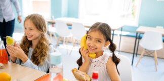 Two smiling girls enjoying snacks during break time in a school classroom.