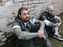 Two tattooed men in prison uniforms sit against a brick wall, exuding a gritty and rebellious vibe.