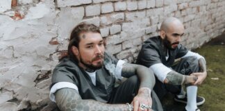 Two tattooed men in prison uniforms sit against a brick wall, exuding a gritty and rebellious vibe.