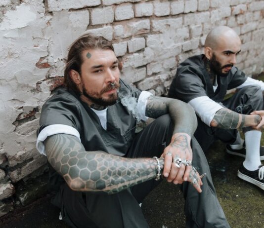 Two tattooed men in prison uniforms sit against a brick wall, exuding a gritty and rebellious vibe.
