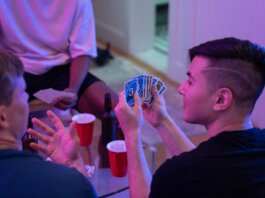 Young adults playing a card game with drinks and laughter in a cozy indoor setting.