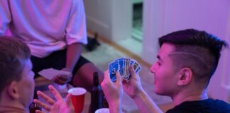 Young adults playing a card game with drinks and laughter in a cozy indoor setting.
