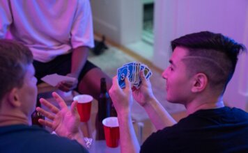 Young adults playing a card game with drinks and laughter in a cozy indoor setting.