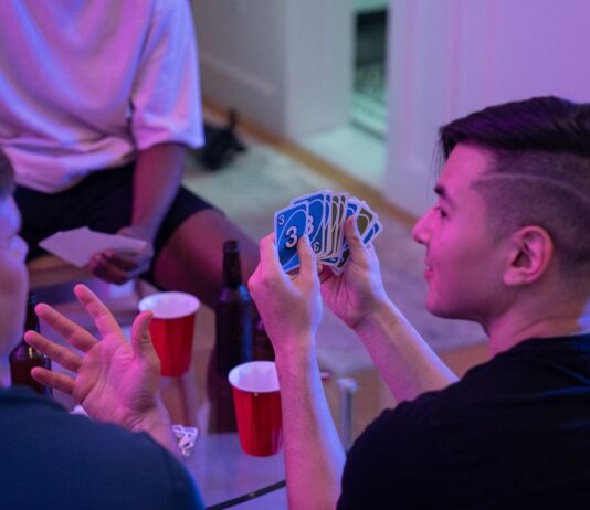 Young adults playing a card game with drinks and laughter in a cozy indoor setting.