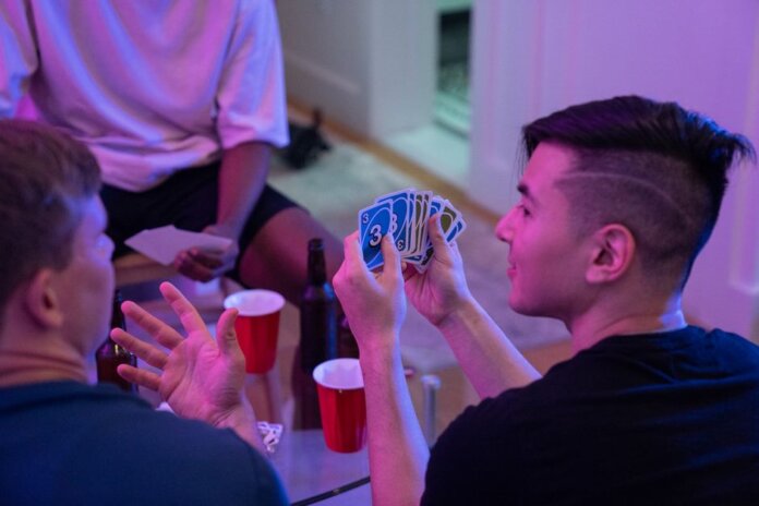 Young adults playing a card game with drinks and laughter in a cozy indoor setting. Young adults playing a card game with drinks and laughter in a cozy indoor setting.
