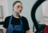 Young woman in a workshop setting, wearing protective goggles and a blue apron, standing indoors.