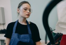 Young woman in a workshop setting, wearing protective goggles and a blue apron, standing indoors.