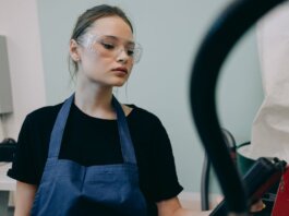 Young woman in a workshop setting, wearing protective goggles and a blue apron, standing indoors.