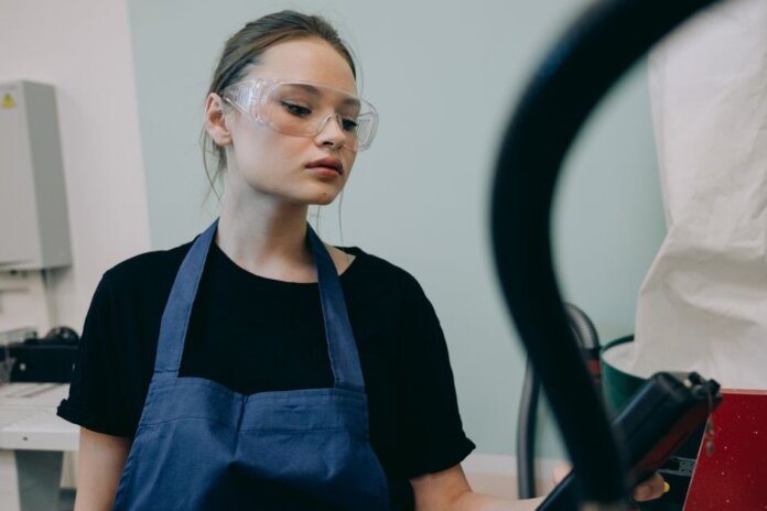 Young woman in a workshop setting, wearing protective goggles and a blue apron, standing indoors. Young woman in a workshop setting, wearing protective goggles and a blue apron, standing indoors.