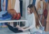 Young woman managing a fashion boutique, multitasking with phone and laptop, surrounded by clothing.