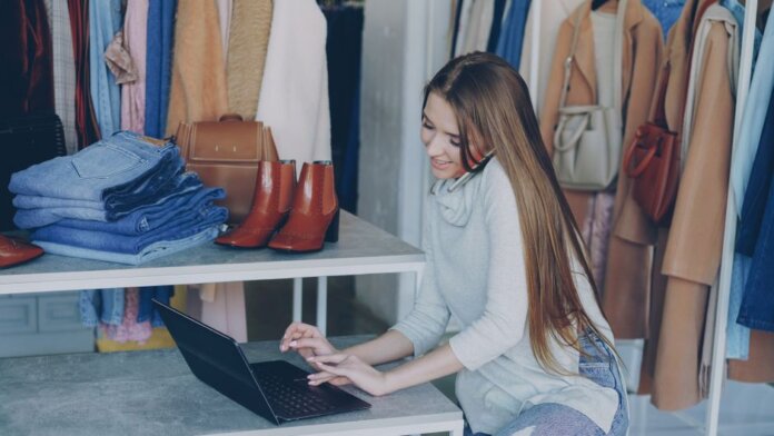 Young woman managing a fashion boutique, multitasking with phone and laptop, surrounded by clothing.