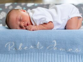 A baby sleeps peacefully on a blue knit blanket with name embroidery.