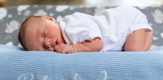 A baby sleeps peacefully on a blue knit blanket with name embroidery.
