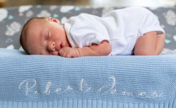 A baby sleeps peacefully on a blue knit blanket with name embroidery.