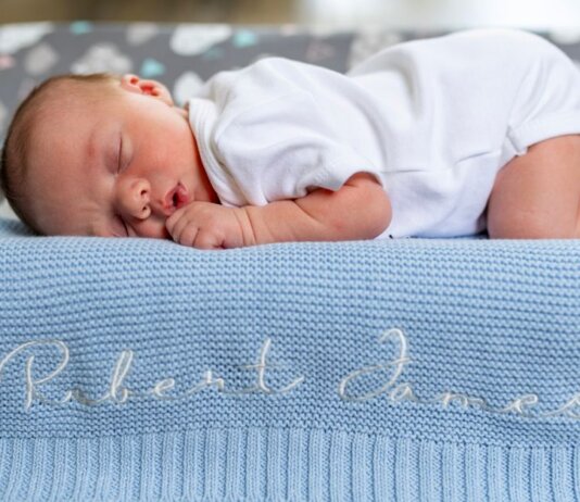 A baby sleeps peacefully on a blue knit blanket with name embroidery.