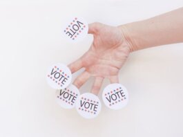 A hand displays several 'Vote' stickers on a white background, symbolizing political participation.