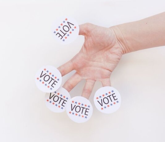 A hand displays several 'Vote' stickers on a white background, symbolizing political participation.