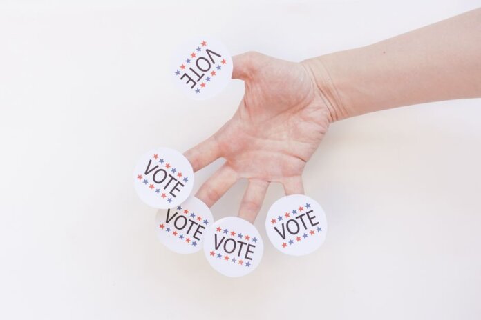 A hand displays several 'Vote' stickers on a white background, symbolizing political participation. A hand displays several 'Vote' stickers on a white background, symbolizing political participation.