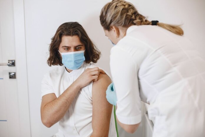 A healthcare worker wearing a mask gives a vaccine shot to a patient in a medical office. A healthcare worker wearing a mask gives a vaccine shot to a patient in a medical office.