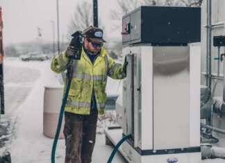 A man wearing a jacket refuels a truck at a gas station during winter with snow falling.