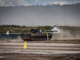 A military tank firing a shell during training in an open field.