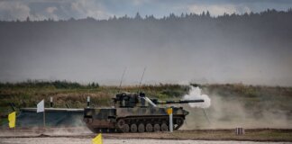 A military tank firing a shell during training in an open field.
