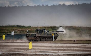 A military tank firing a shell during training in an open field.