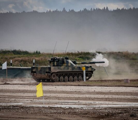 A military tank firing a shell during training in an open field.
