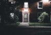 A warmly lit brick house entrance under a night sky, featuring a serene garden setting.