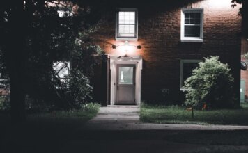A warmly lit brick house entrance under a night sky, featuring a serene garden setting.