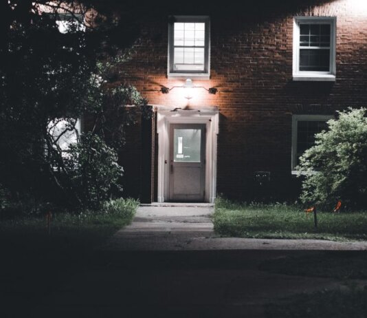 A warmly lit brick house entrance under a night sky, featuring a serene garden setting.