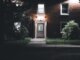 A warmly lit brick house entrance under a night sky, featuring a serene garden setting.