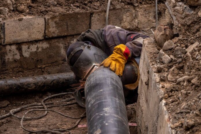 A worker welds a pipe in an urban trench, showcasing industrial construction work. A worker welds a pipe in an urban trench, showcasing industrial construction work.